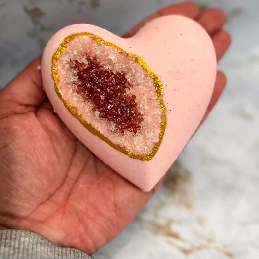 Heart-shaped bath bomb with a geode-like center held in a hand against a marble background