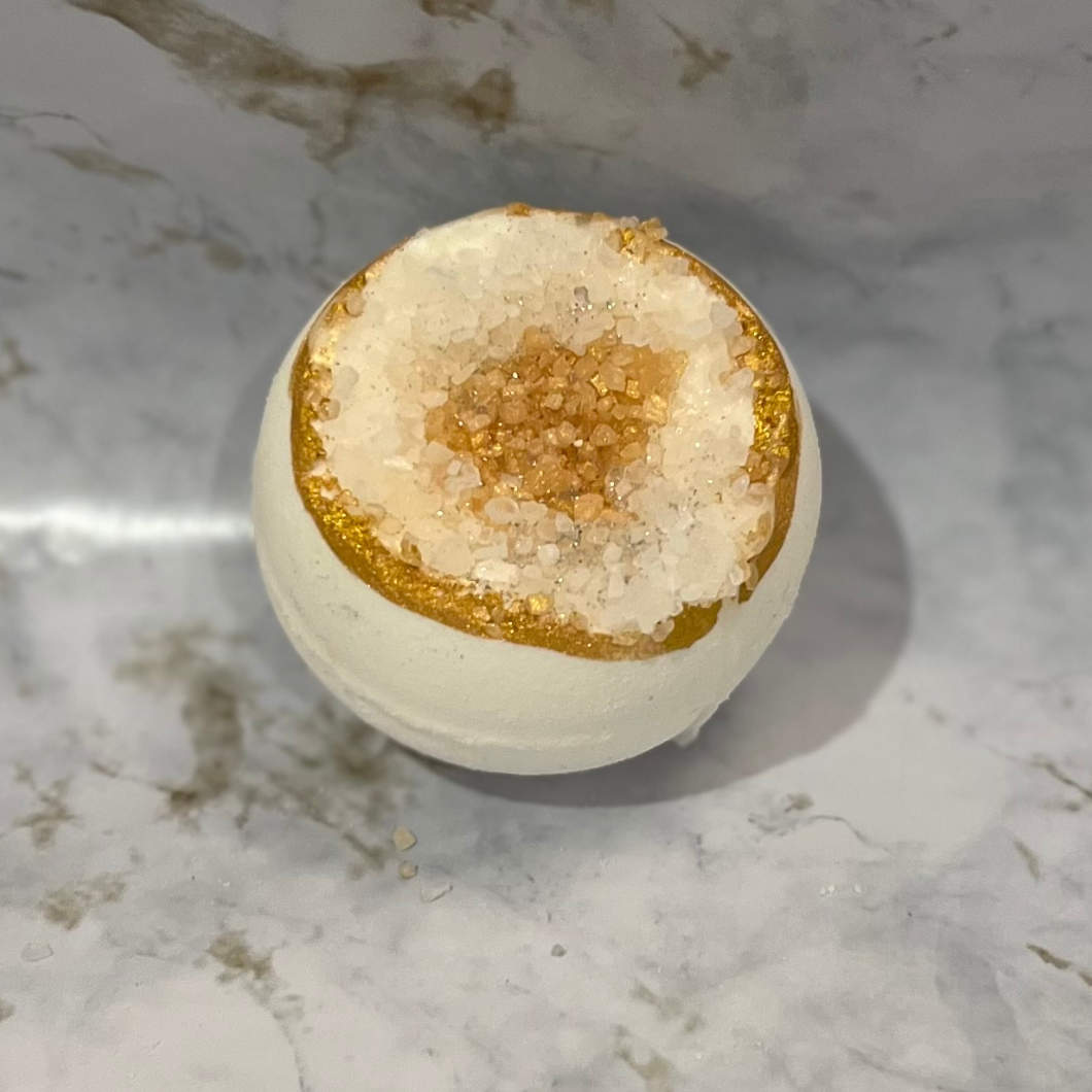 A bath bomb with a geode-inspired design, featuring a gold citrine-like structure on top with clear and gold hues, placed on a marble surface.