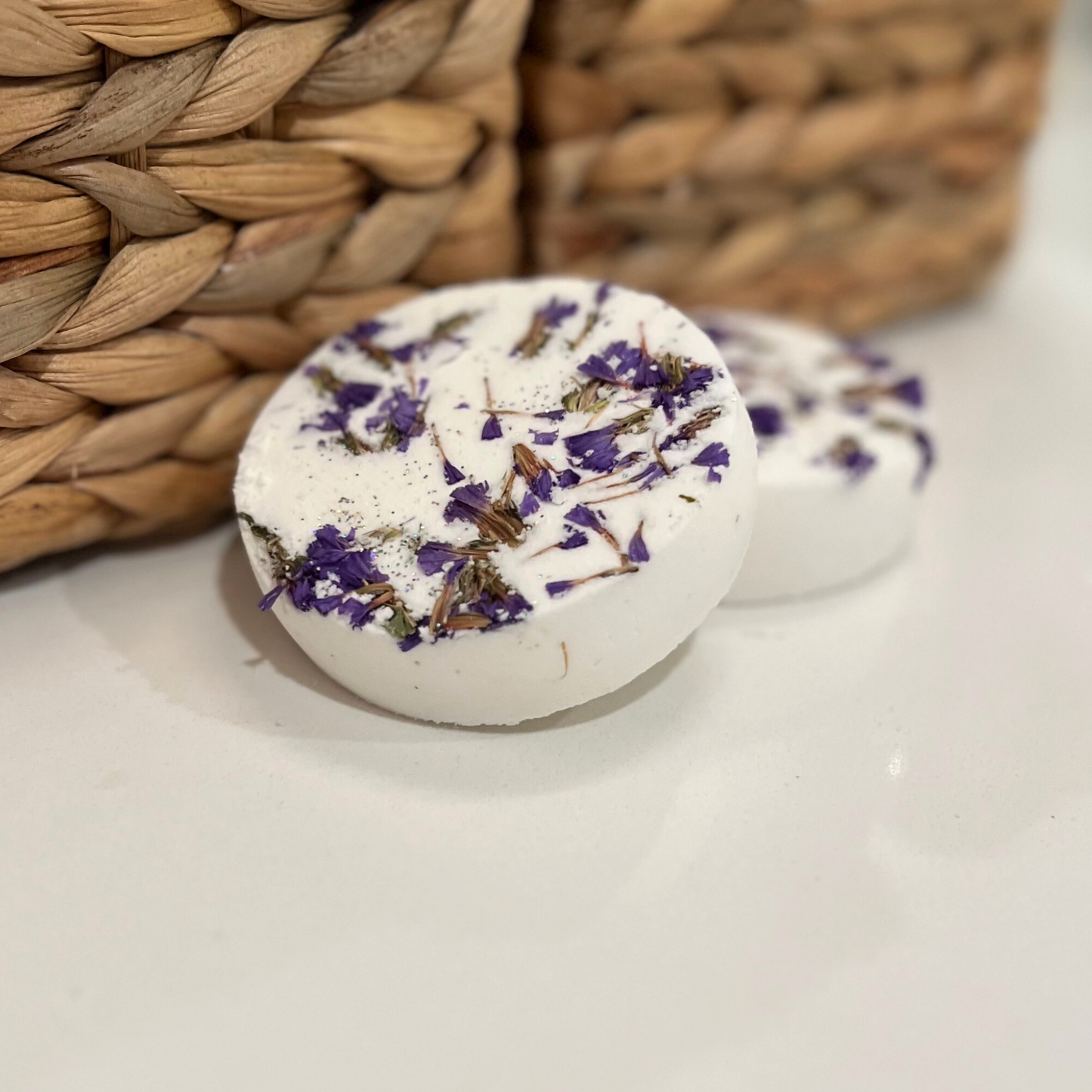 Two shower steamers in white with purple flowers displayed next to a woven background 