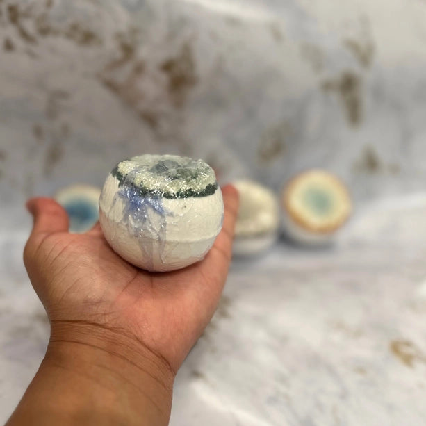 Hand holding a white bath bomb with more bath bombs in the background on a marble surface.