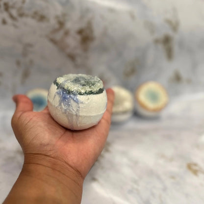 Hand holding a white bath bomb with more bath bombs in the background on a marble surface.