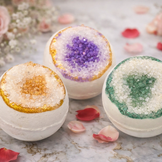 A close up of three white bath bombs, one with a purple center, one with a gold center and one with a gree center, arranged on a marble surface with flowers surrounding them.
