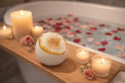 One white bath bomb with a gold center, arranged on a brown bath tray with a candle beside it and a tub with rose petals in the background.