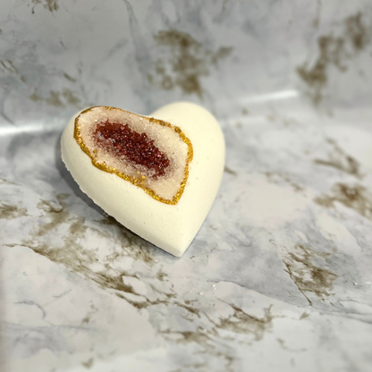 A heart-shaped bath bomb with a geode design, featuring a central pocket of red sea salt, with a white exterior and gold mica accents.