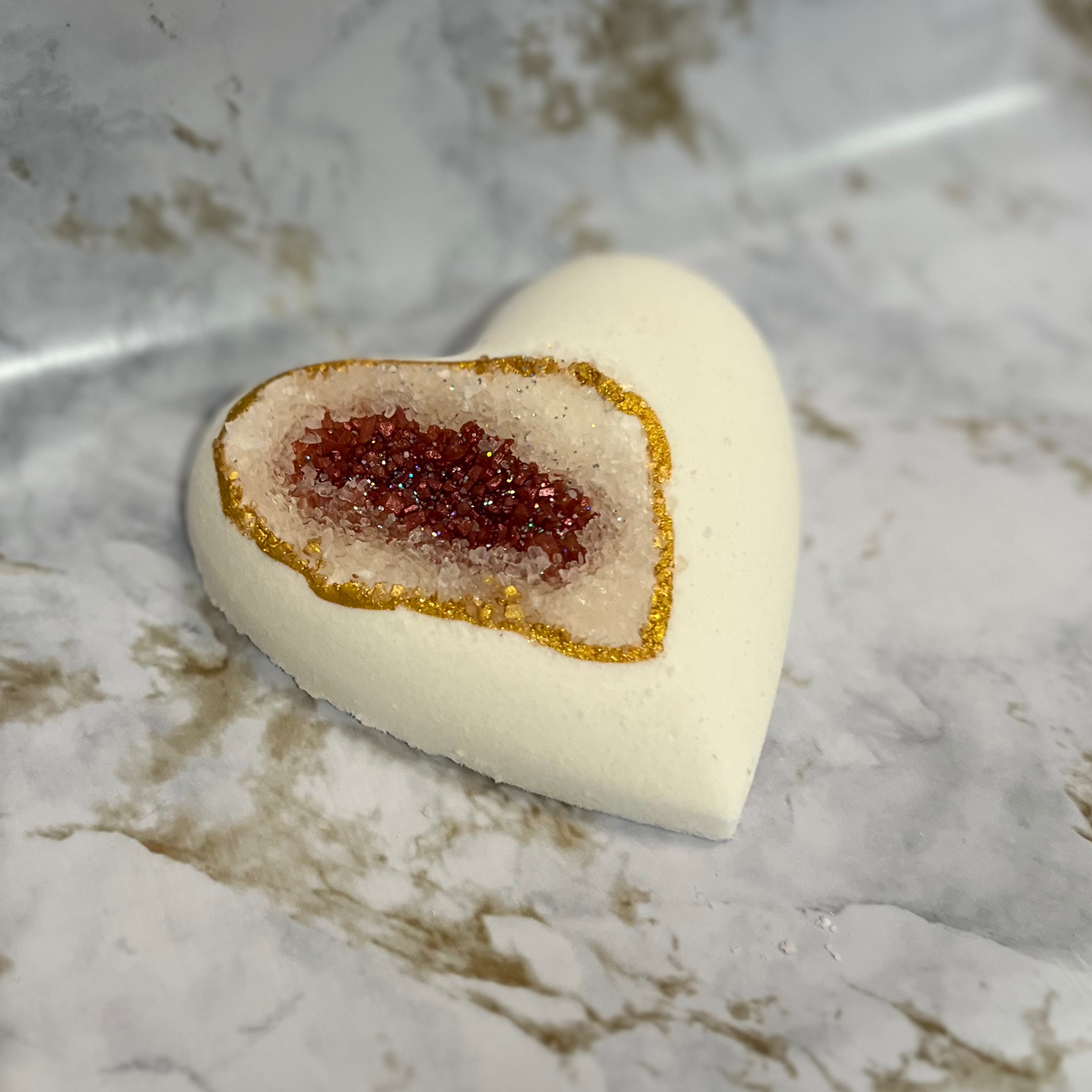A heart-shaped bath bomb with a geode design, featuring a central pocket of red sea salt, with a white exterior and gold mica accents.