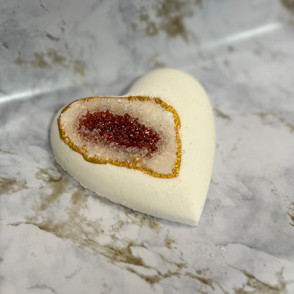 A heart-shaped bath bomb with a geode design, featuring a central pocket of red sea salt, with a white exterior and gold mica accents.