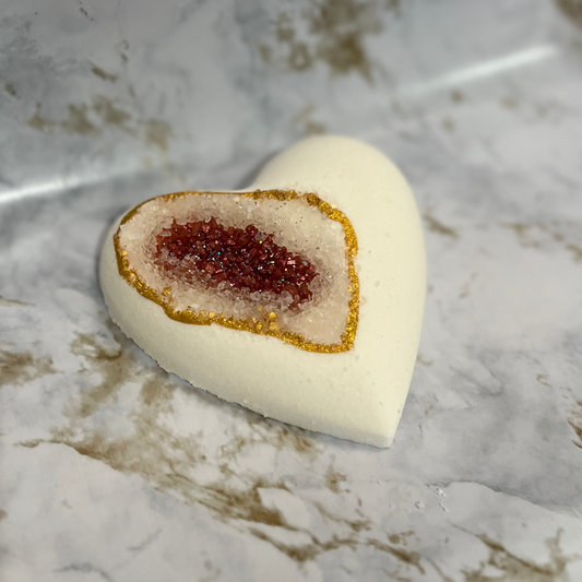A heart-shaped bath bomb with a geode design, featuring a central pocket of red sea salt, with a white exterior and gold mica accents.