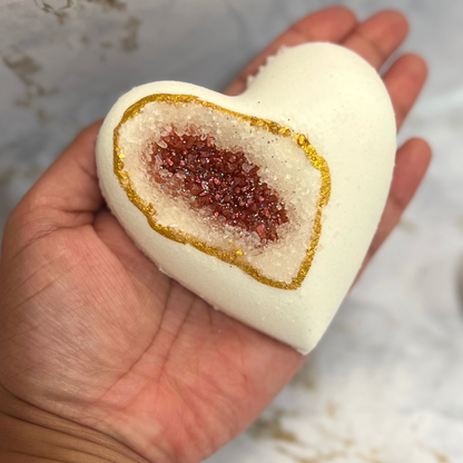 A hand holding a heart-shaped bath bomb with a geode design, featuring a central pocket of red sea salt, with a white exterior and gold mica accents.