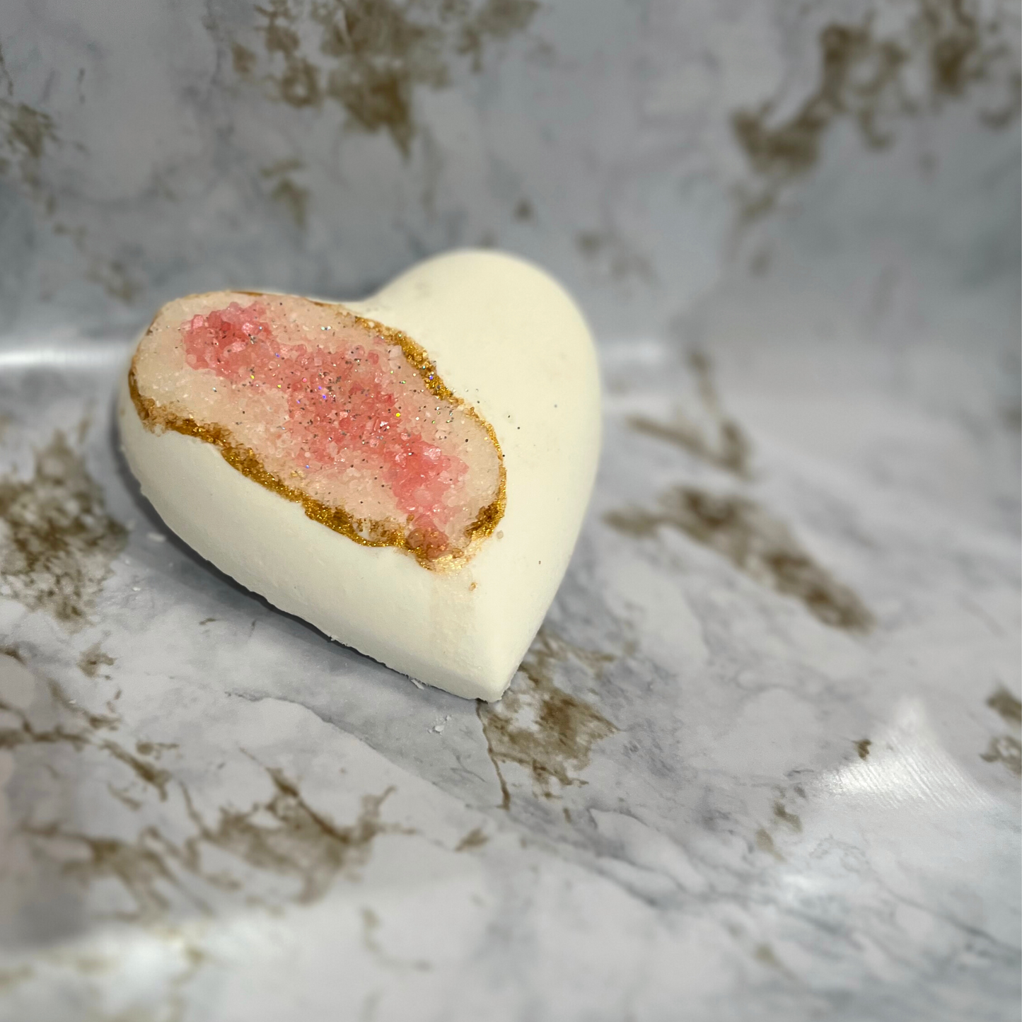 A heart-shaped bath bomb with a geode-like pink center and white exterior, adorned with biodegradable glitter along the edge.