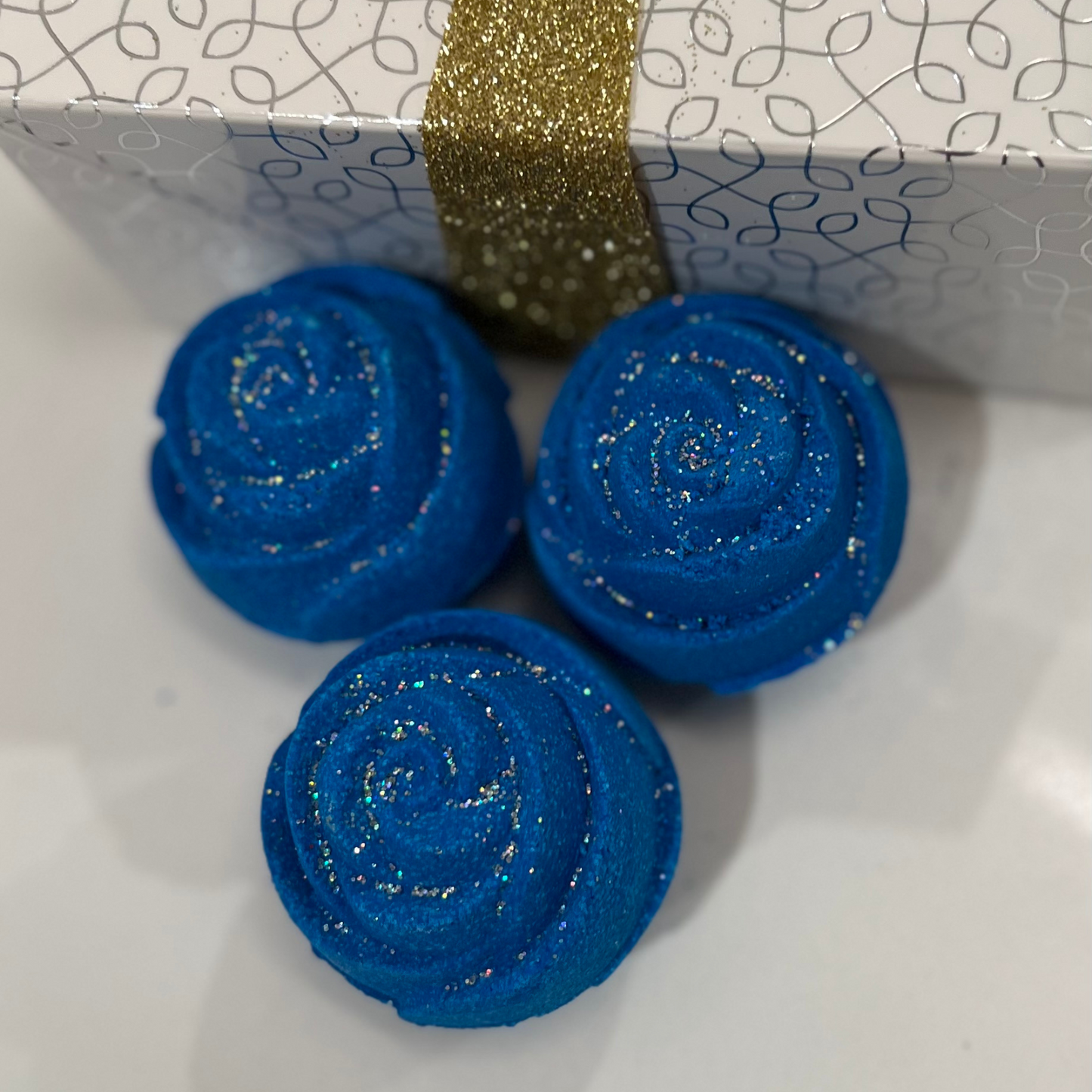 Three blue rose-shaped bath bomb with glitter, against a white box with a silver design and gold glitter ribbon.