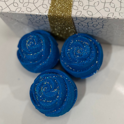 Three blue rose-shaped bath bomb with glitter, against a white box with a silver design and gold glitter ribbon.
