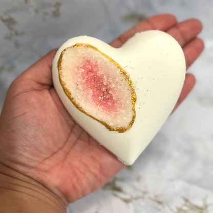 A hand holding a heart-shaped bath bomb with a geode-like pink center and white exterior, adorned with biodegradable glitter along the edge.