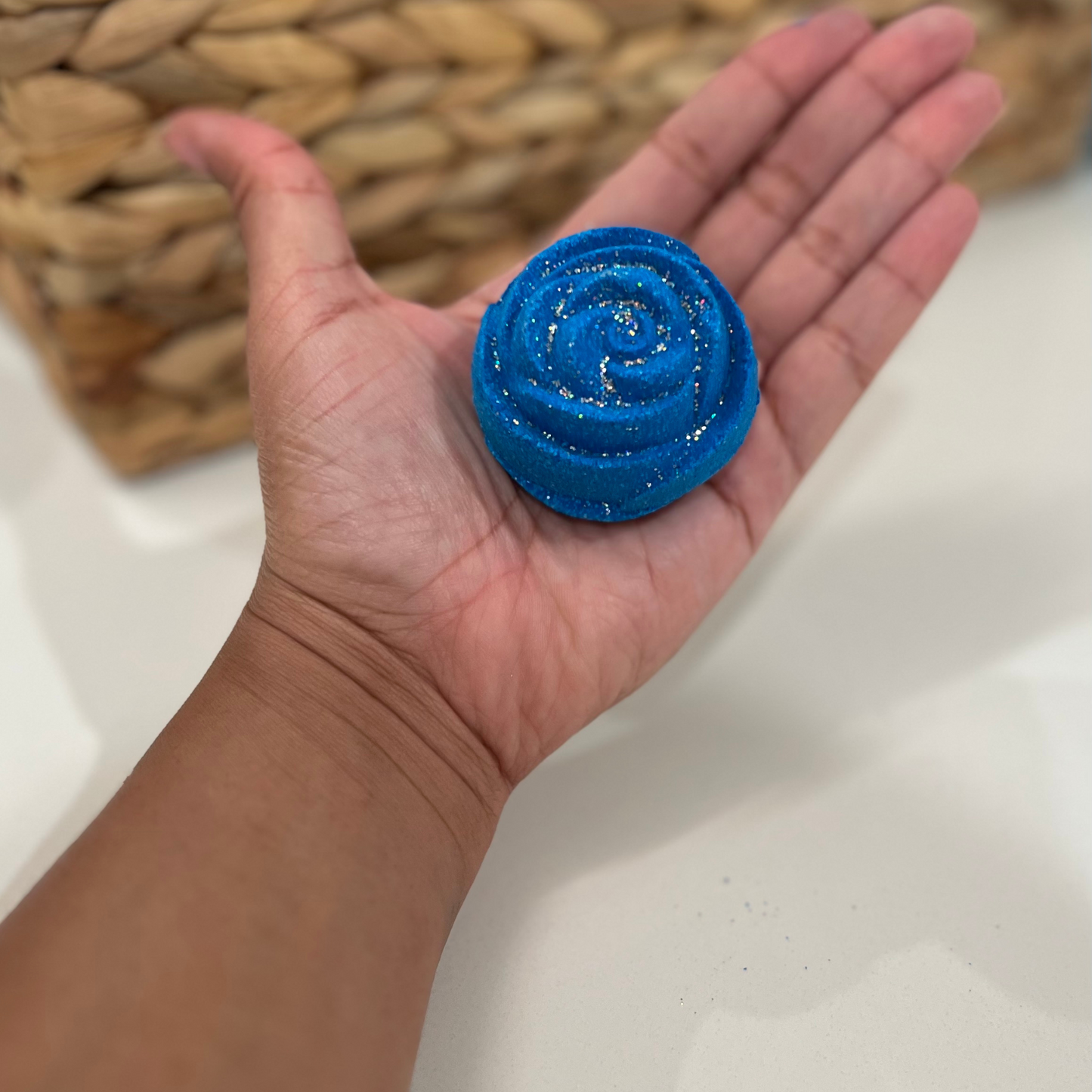 A hand holding a blue rose-shaped bath bomb with glitter, against a woven background.