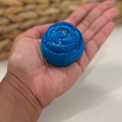 A hand holding a blue rose-shaped bath bomb with glitter, against a woven background.