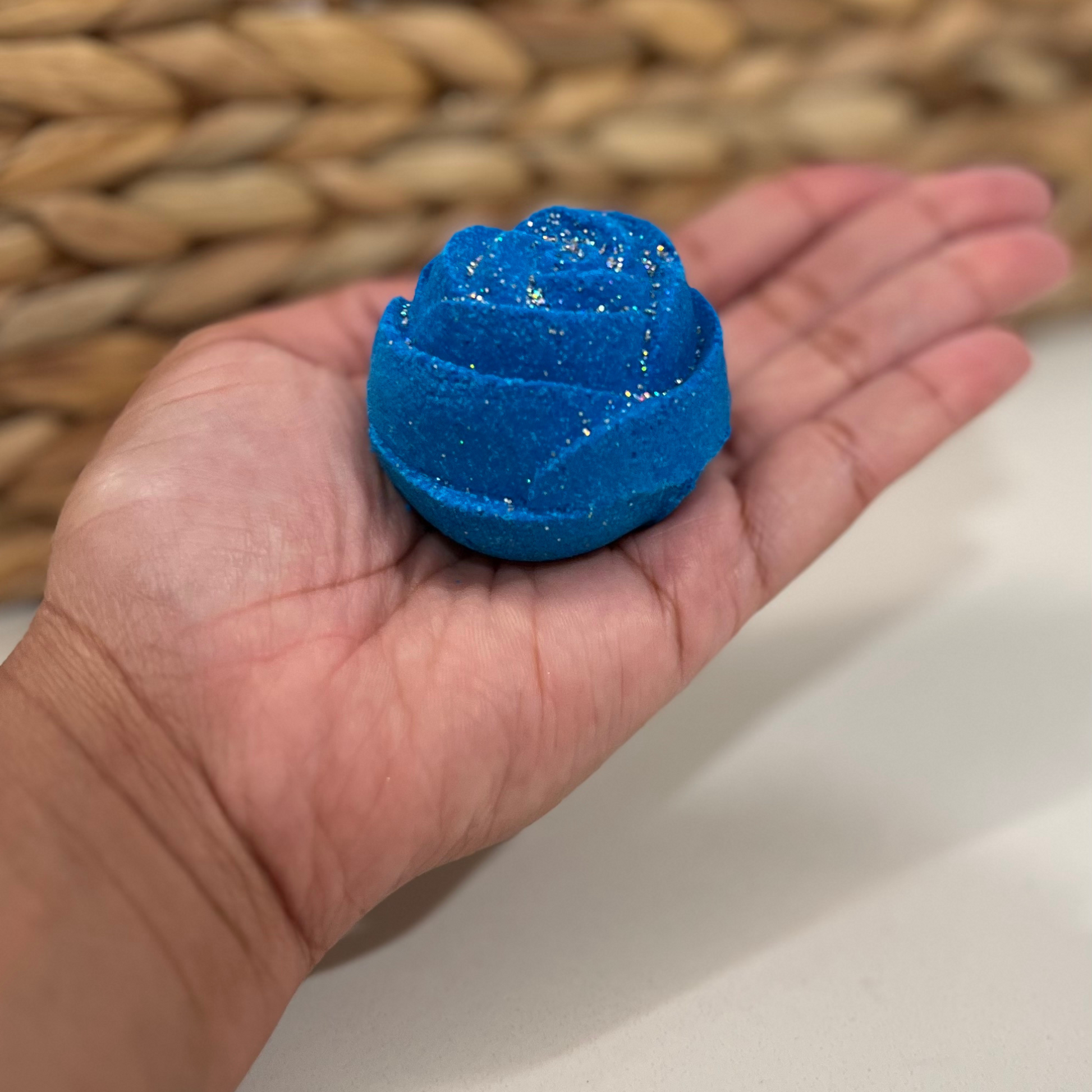 A hand holding a blue rose-shaped bath bomb with glitter, against a woven background.