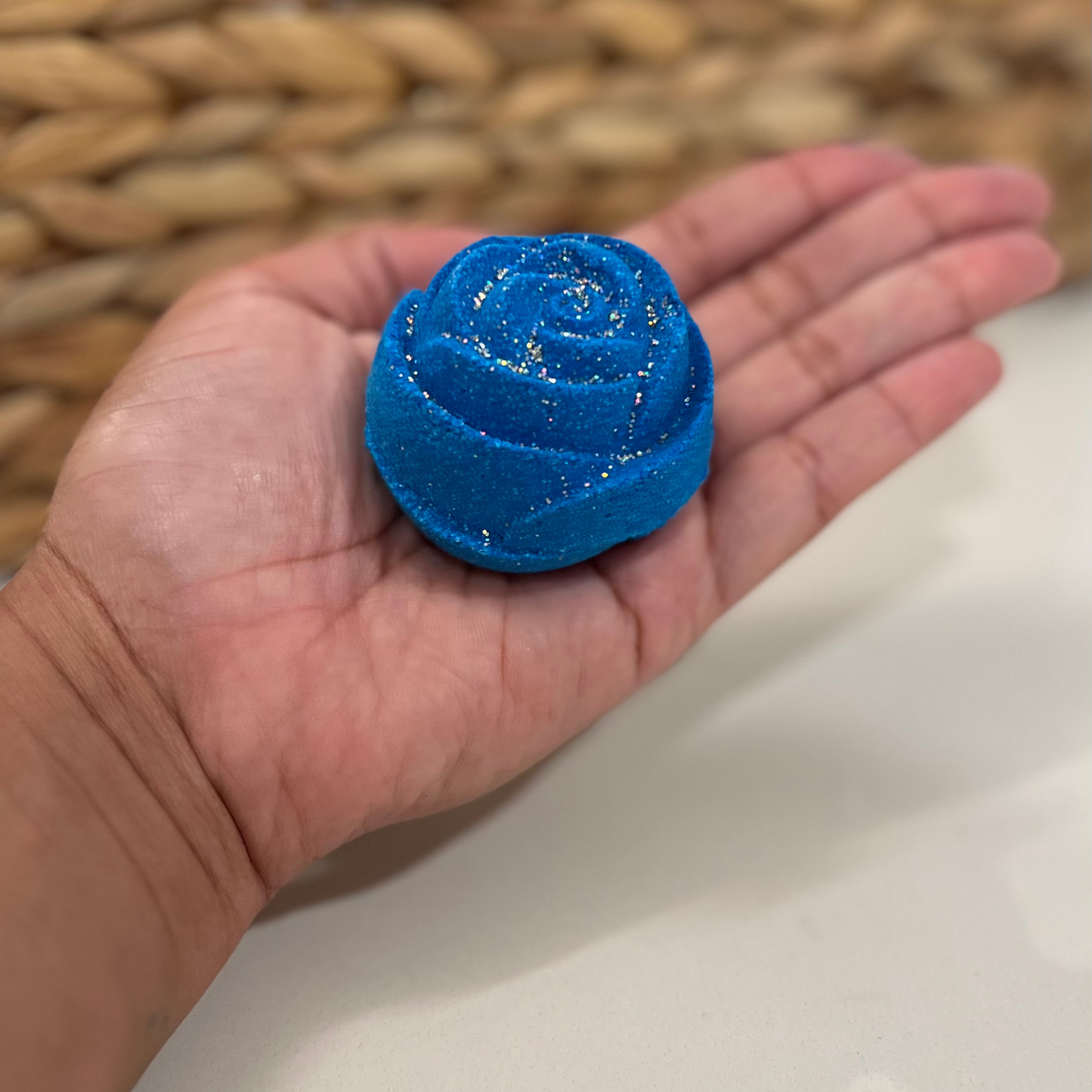 A hand holding a blue rose-shaped bath bomb with glitter, against a woven background.