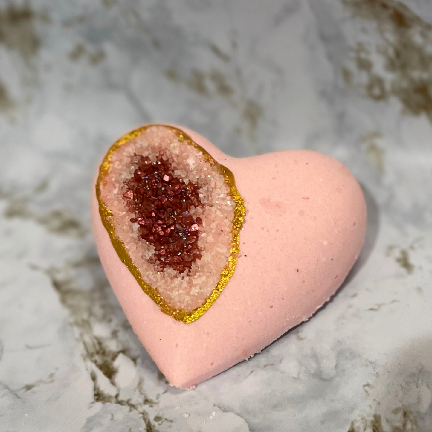 A heart-shaped bath bomb with a geode pattern, featuring a pink exterior and a central dark red sea salt. The bath bomb is adorned with biodegradable glitter along its edge.