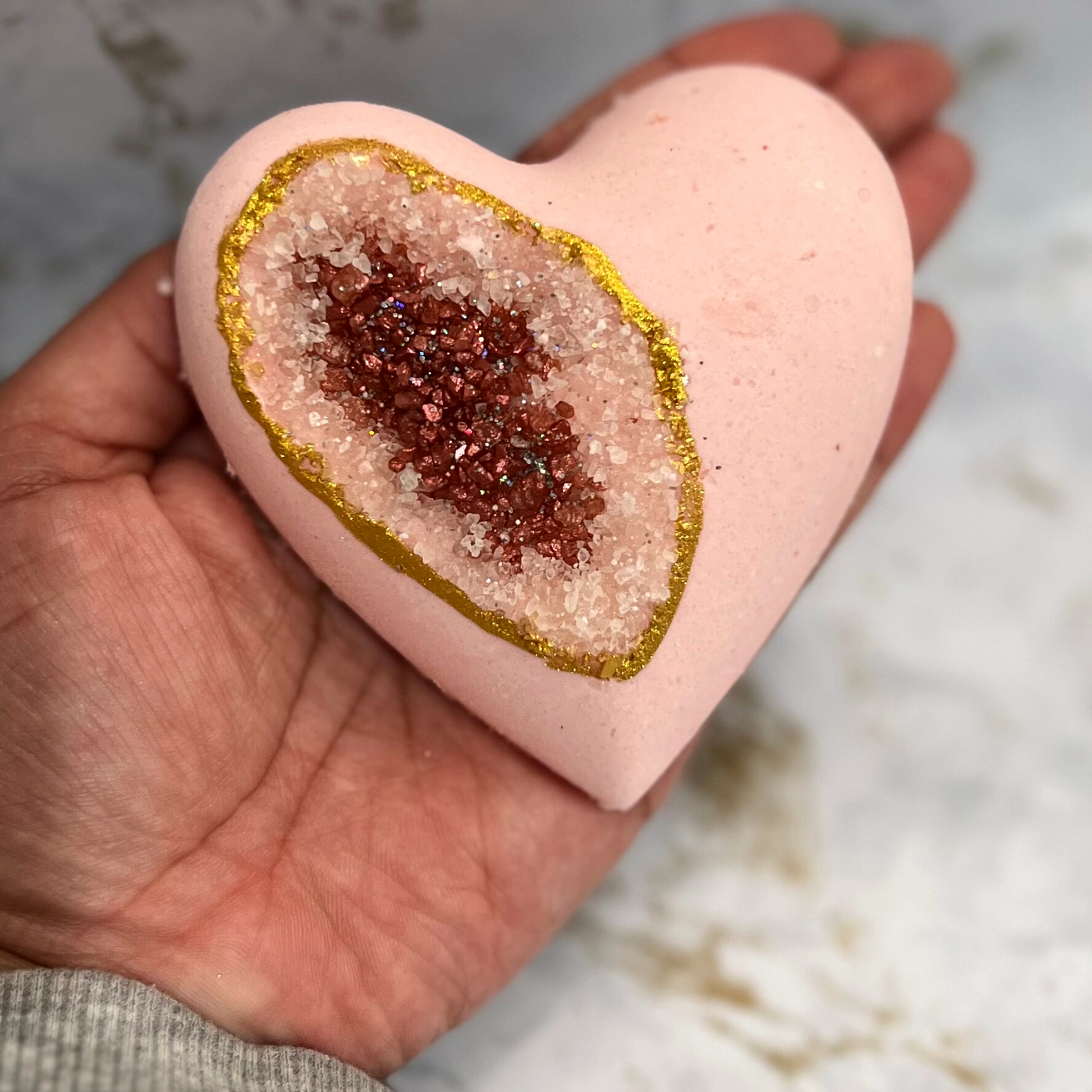 A hand holding a heart-shaped bath bomb with a geode pattern, featuring a pink exterior and a central dark red sea salt. The bath bomb is adorned with biodegradable glitter along its edge.