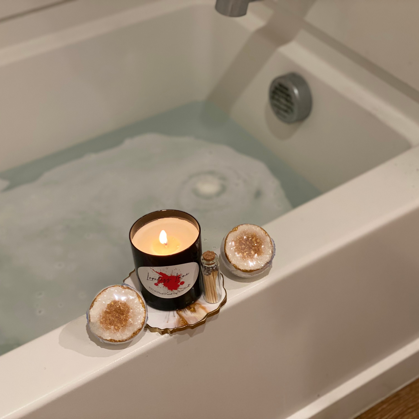 A bath tub with two bath bomsb with a geode-inspired design, featuring a gold citrine-like structure on top with clear and gold hues, and a lit candle on a gold and white marble coaster with a set of matches next to it, placed on the edge of the tub.