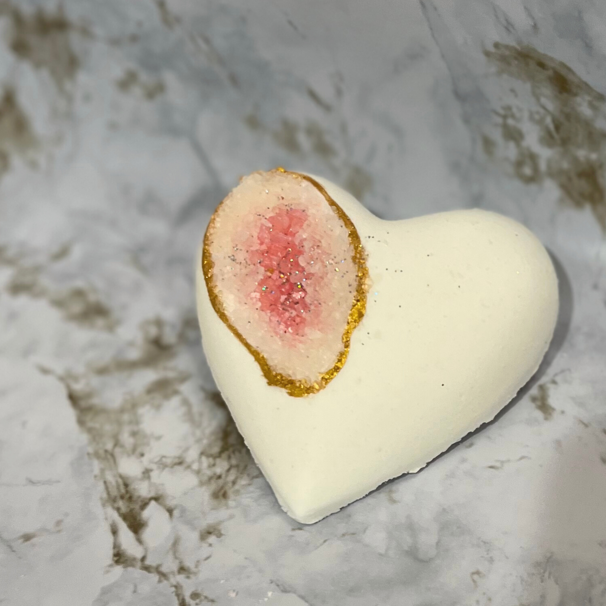 A heart-shaped bath bomb with a geode-like pink center and white exterior, adorned with biodegradable glitter along the edge.